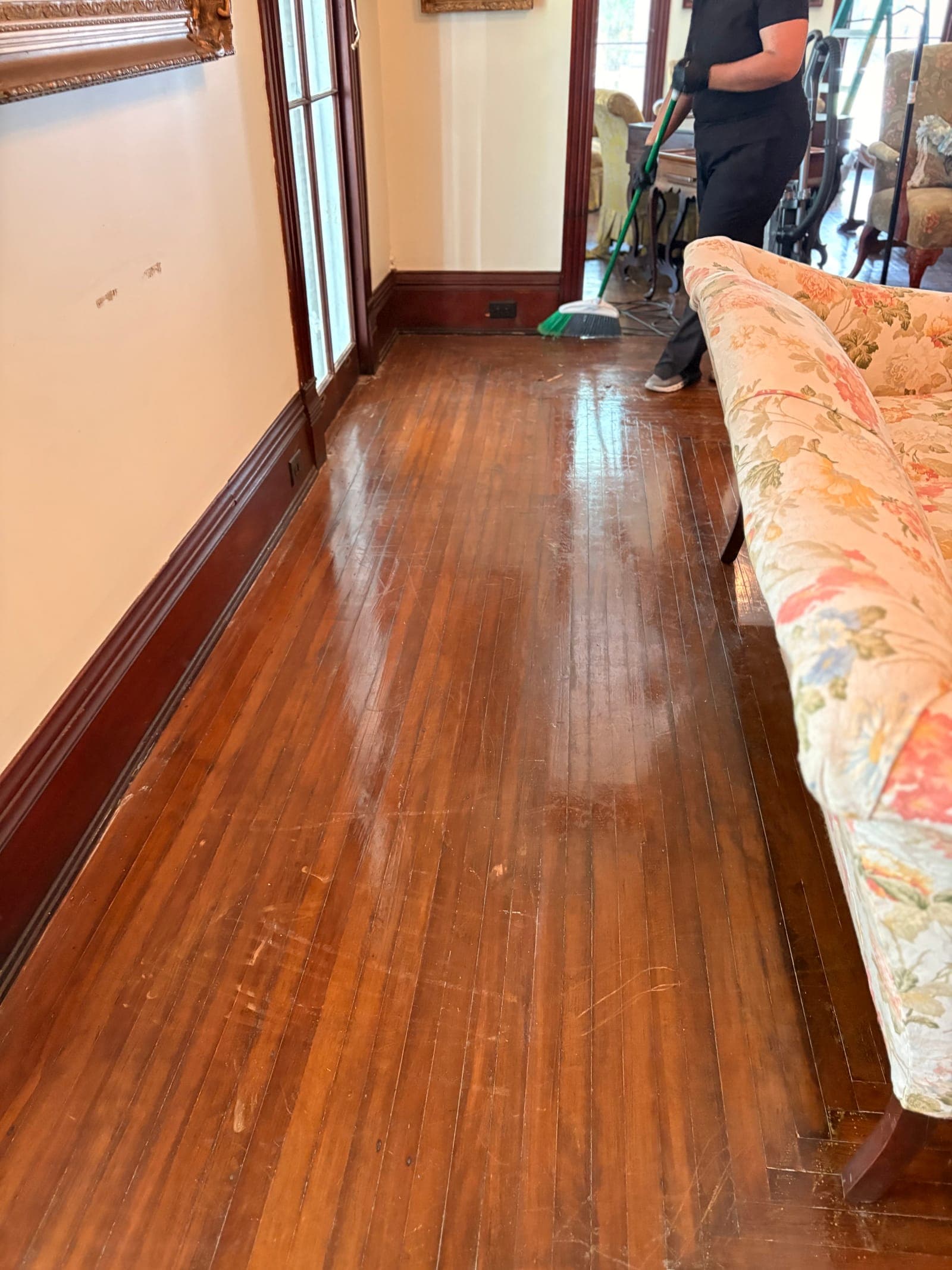 A CC Cleaning Solutions team member sweeping a hardwood hallway beside a floral sofa
